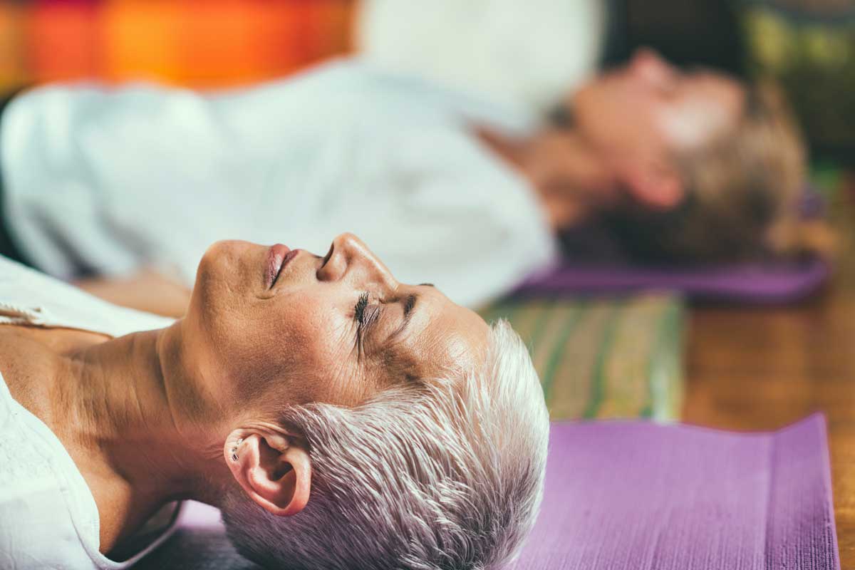 Close-up shot of two individuals lying on their backs on yoga mats, engaging in a mindfulness meditation session. They are relaxed, eyes closed, focusing on their breathing and listening to a meditation mantra. The background is softly blurred, emphasizing the calm and peaceful atmosphere.