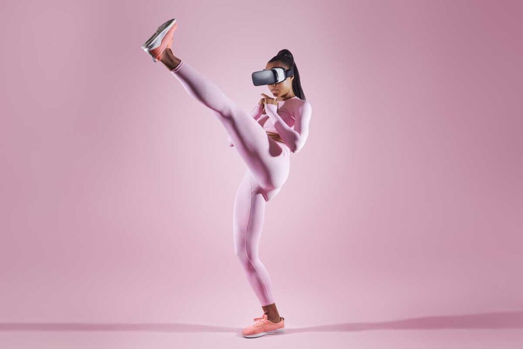 Woman in virtual reality headset practicing in kicking while standing against pink background.