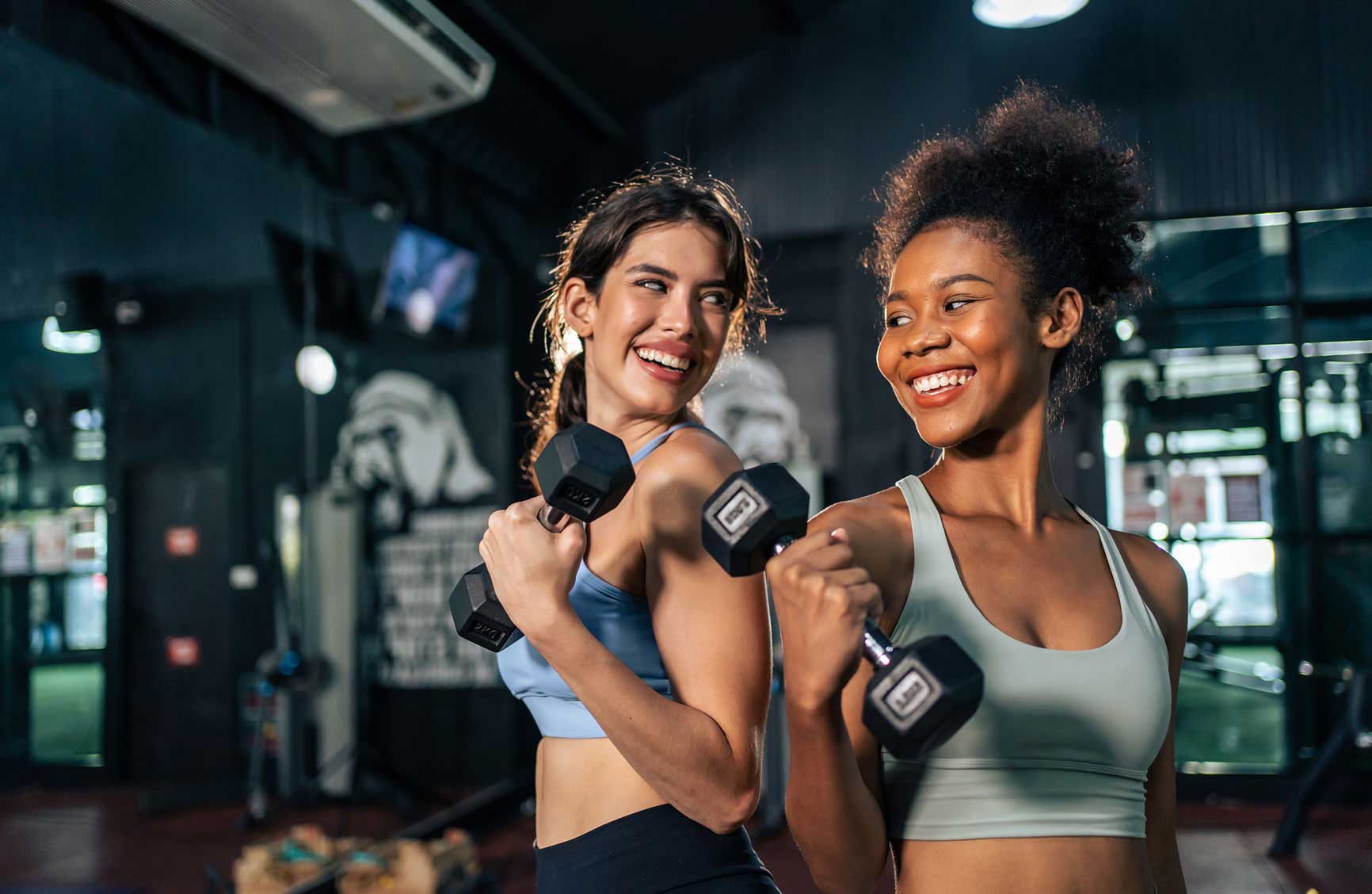 Two women smiling and lifting dumbbells during a fitness workout session in a gym.