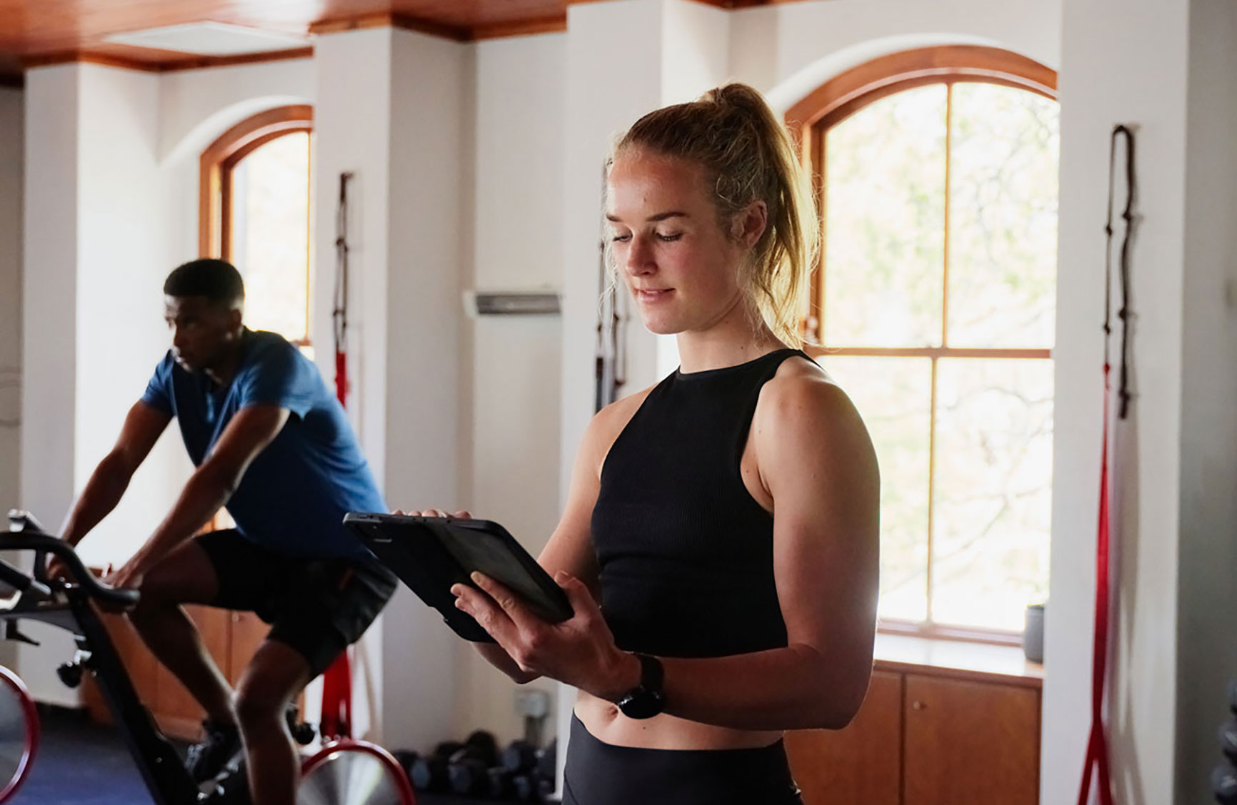 Woman in a fitness studio using a tablet, with a man cycling in the background.