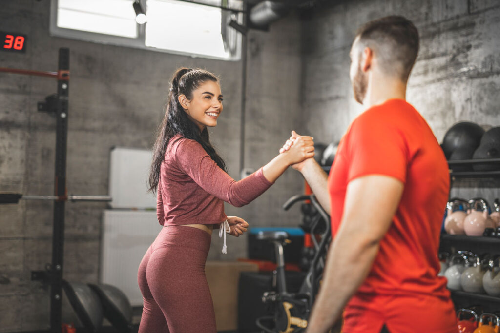 Woman in a gym giving a handshake to a man in a red shirt, both smiling after a workout session.