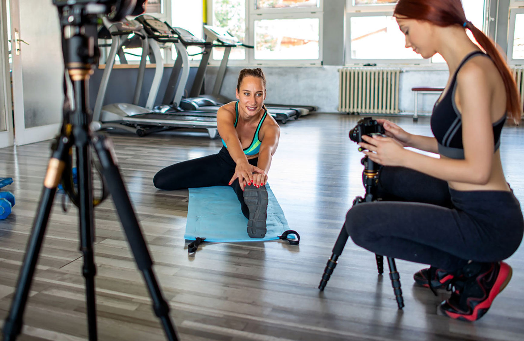 A fitness trainer stretching on a mat while being filmed by a videographer in a gym.